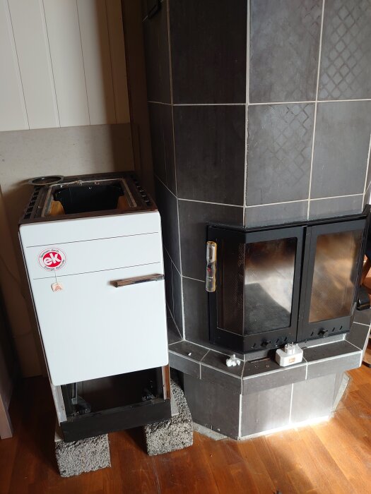 Ny vedspis på plintar bredvid modern mörkgrå tegelkamin i ett hem, redo för installation.