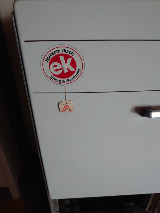 En vit apparat med en röd etikett som säger "Sparsam durch Energie-Kontrolle" och en liten hängande skylt, bredvid en täljstensspis.