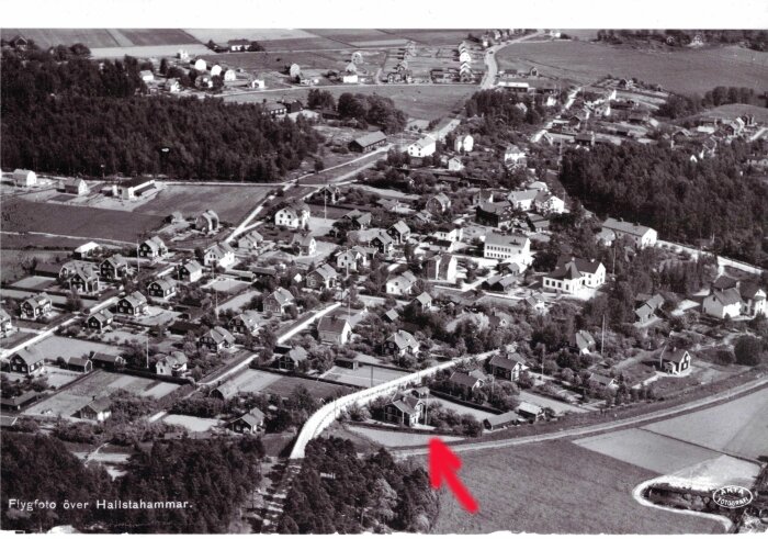 Flygfoto över Hallstahammar med en röd pil som pekar på ett specifikt hus, omgiven av flera andra byggnader och en skog i bakgrunden.