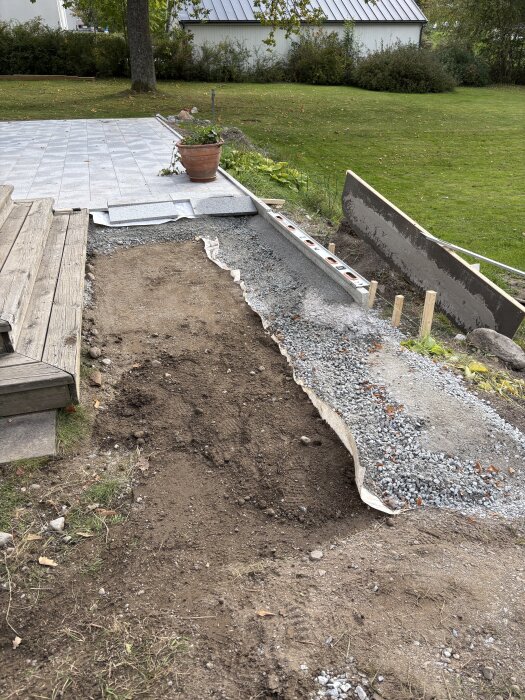 Grusigt område vid sidan av ett trädäck under pågående markarbeten för framtida utekök, med plattor och en blomkruka i bakgrunden.