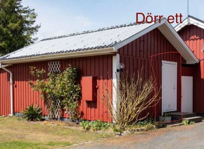 Röd sommarstuga med vit dörr markerad i rött, omgiven av buskar. Taket är vitt och fasaden har rödmålade panelbrädor. Text "Dörr ett" visas.