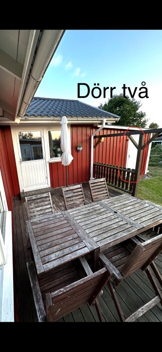Trädäck med träbord och stolar, röd stugfasad i bakgrunden med vit ytterdörr och fönster, samt texten 'Dörr två'.