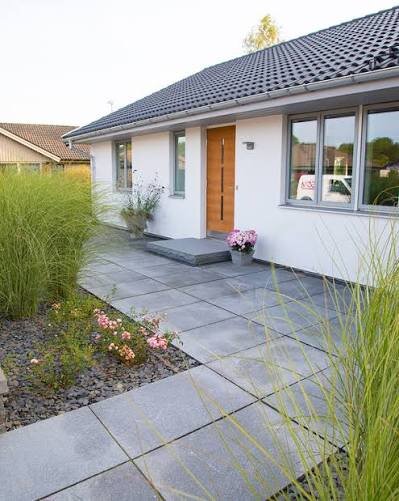 Ingång till ett hus med grå plattläggning, gröna prydnadsgräs och en planterad rabatt bredvid.