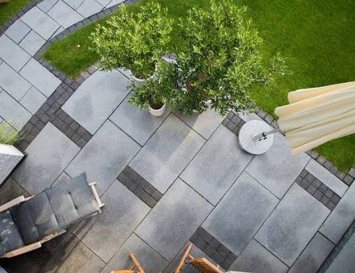 Grå stenterrass med solstol och parasoll, omgiven av grön gräsmatta och träd.