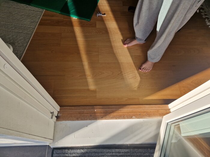 Öppen altandörr med synlig dörrtröskel och spår där dörrlister tidigare suttit. Trägolv och personens bara fötter syns i bild.