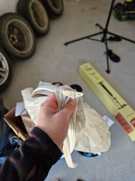 Hand håller upp en vit dörrlist i plast, omgiven av olika föremål i en garage- eller förrådsmiljö.