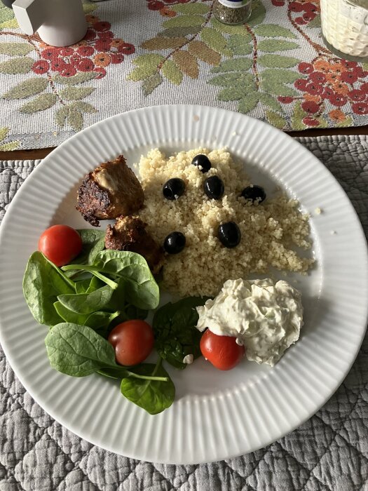 Tallrik med fiskbullar, couscous, oliver, spenat, tomat och yoghurtsås på bord med blommig duk.