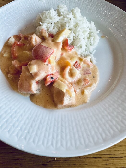 Fisk- och skaldjursgryta med ris på vit tallrik, innehållande räkor och kräftor, i en krämig sås med bitar av grönsaker.