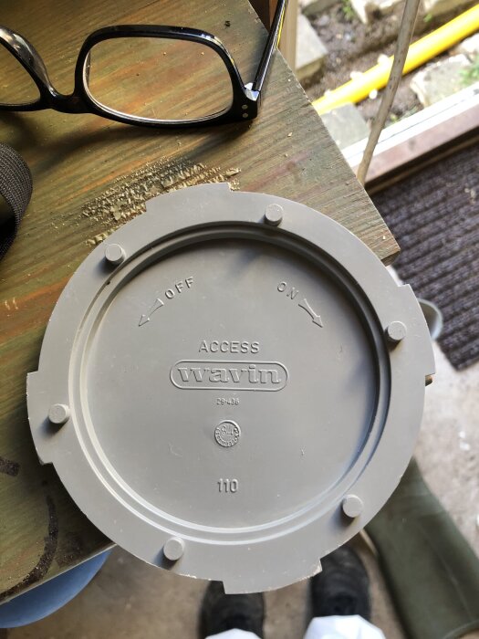 Grått plastlock från "wavin" med texten "ACCESS" och "110", liggande på träyta bredvid glasögon.