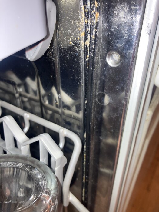 Närbild på smuts och matrester samlade i packningen mellan diskmaskinens diskutrymme och metallfront. En glasburk syns också.