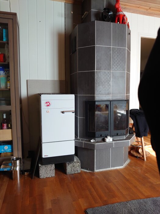 En vit spis står provisoriskt på betongblock bredvid en mörk kakelbeklädd spis i ett rum med trägolv och panelklädda väggar.