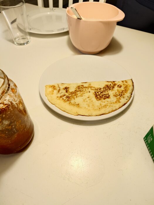 En tallrik med en pannkaka på ett bord, bredvid en rosa bunke och en burk sylt.
