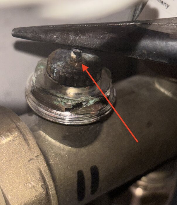 Ärgad ventil med pigg visas, tång trycker ner piggen, möjligt tecken på läckage syns som liten droppe. Ventilen är del av golvvärmesystem.