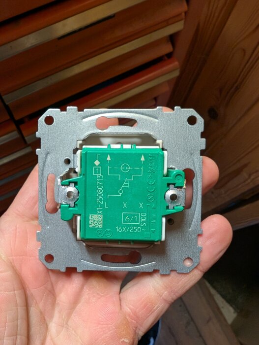 En hand håller en elektrisk brytare med gröna och metalliska delar, där symboler och texter finns utskrivna på brytaren.