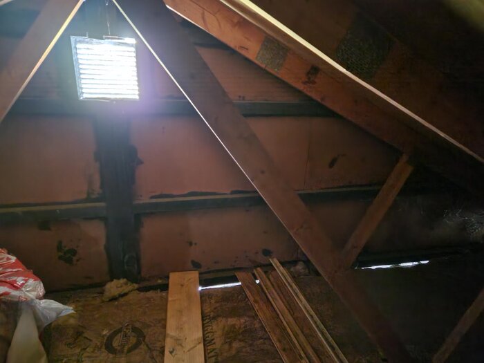 Vind med hängande plywood ovanför och träbjälkar; synlig ventilationsöppning och brädor på golvet i byggprojekt.