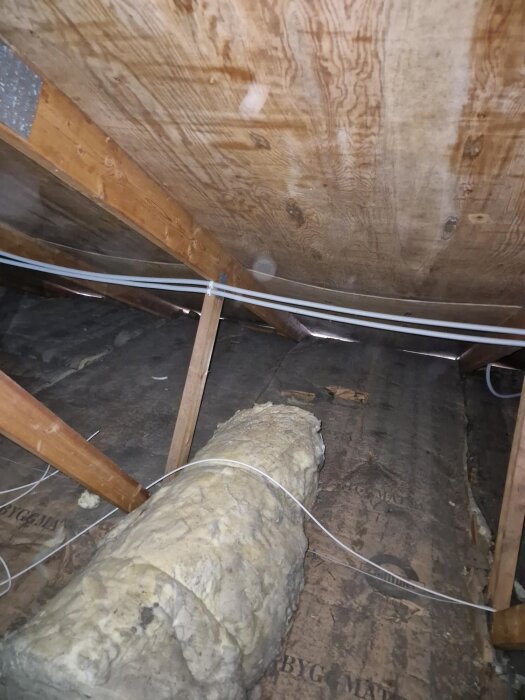 Träbjälkar och hängande plywood i taket på en vind, med isoleringsmaterial och kablar. Ventilationsfråga diskuteras i sammanhanget.