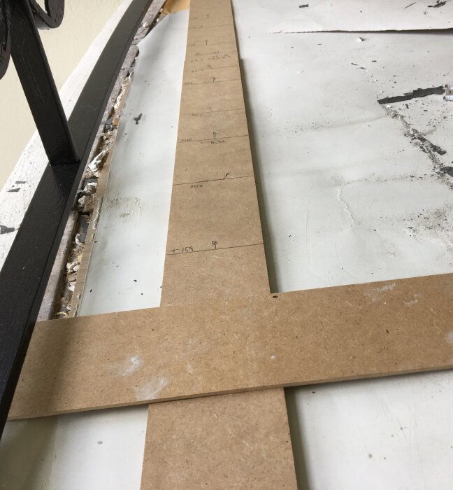 Mätmall av MDF-skiva placerad mot en mur och smidesräcke, med handskrivna mått för golvlistmontering.