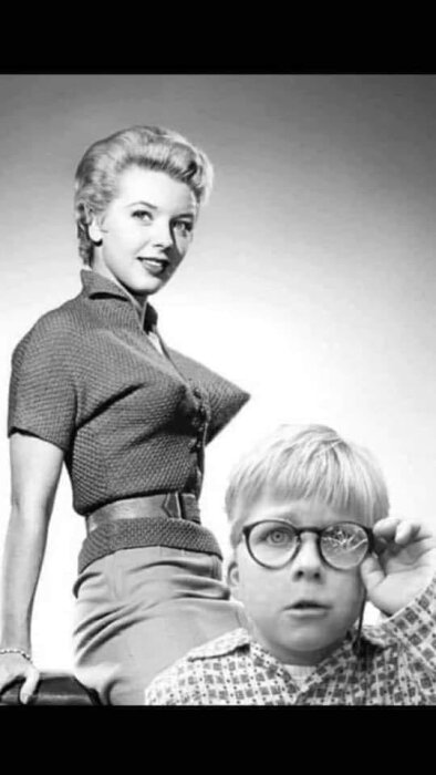 Svartvit bild av en kvinna i en tröja med spetsig detalj och ett barn med glasögon som tittar förvånat genom en förstoringsglaslins.