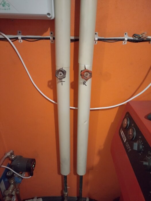 Två vita rör med ventiler märkta "T" och "R" mot en orange vägg bredvid en röd värmepanna.