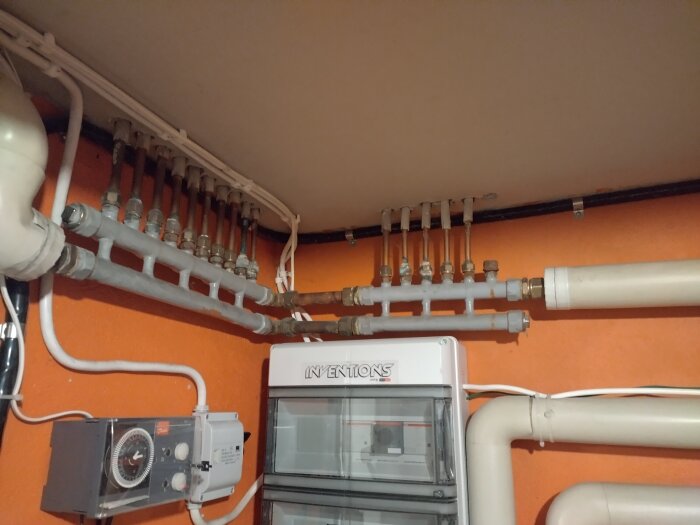 Värmesystem med rör och kopplingar monterade mot en orange vägg, del av uppvärmningsinstallation med mätare och kablar synliga.