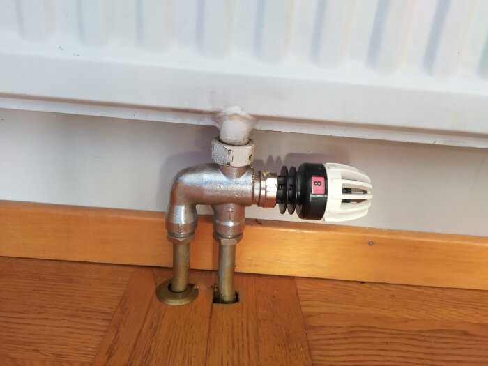 Radiator med termostatventil och två anslutningsrör in i trägolv.