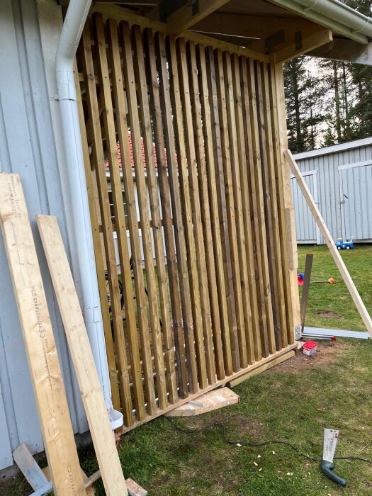 Vertikala ribbor i impregnerat trä monterade på sidan av en carport, bredvid lösa brädor och verktyg på gräsmattan.