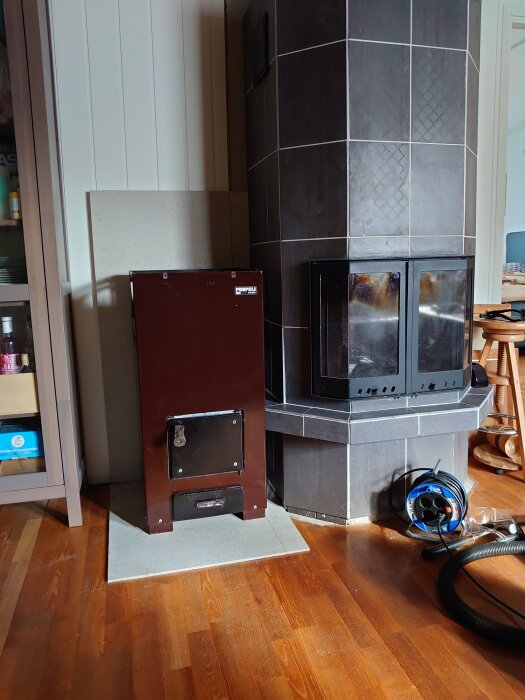 Brun Puupeli vedspis bredvid en grå kakelugn i ett rum med trägolv.