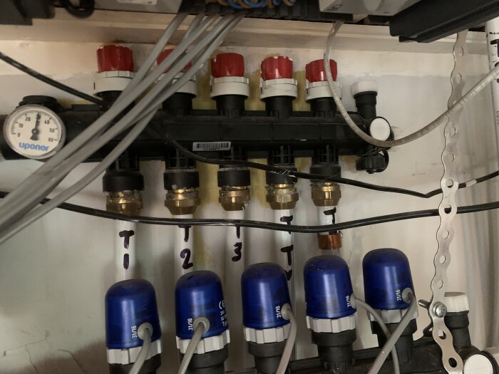 Golvvärmefördelare med fem märka slangar och ventiler, tre är numrerade och markerade med blå ventiler. Kablage och mätare syns även i bilden.