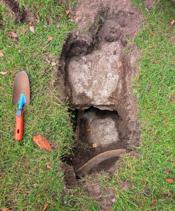 Grop i gräsmatta med en stor platt sten synlig under jord. En spade med orange handtag ligger bredvid. Grön gräsmark runtom.