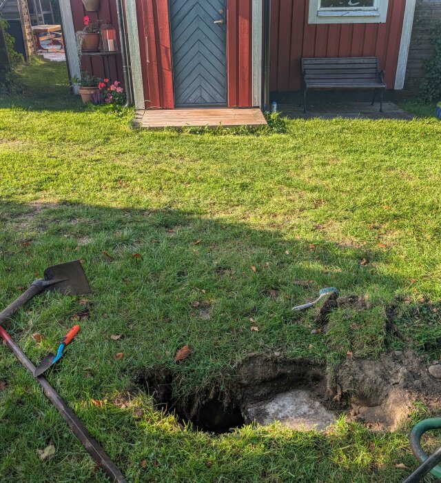Grop i gräsmatta framför röd stuga, synlig platt sten under marken. Trädgårdsverktyg ligger bredvid gropen.‍