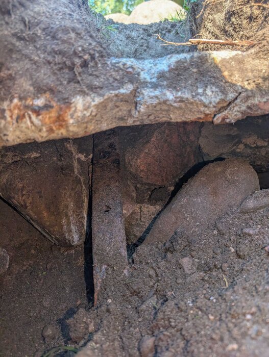 Underjordisk struktur med platt sten och böjt plattjärn synligt från uppgrävd grop nära sommarhus. Omges av mindre stenar och jord.