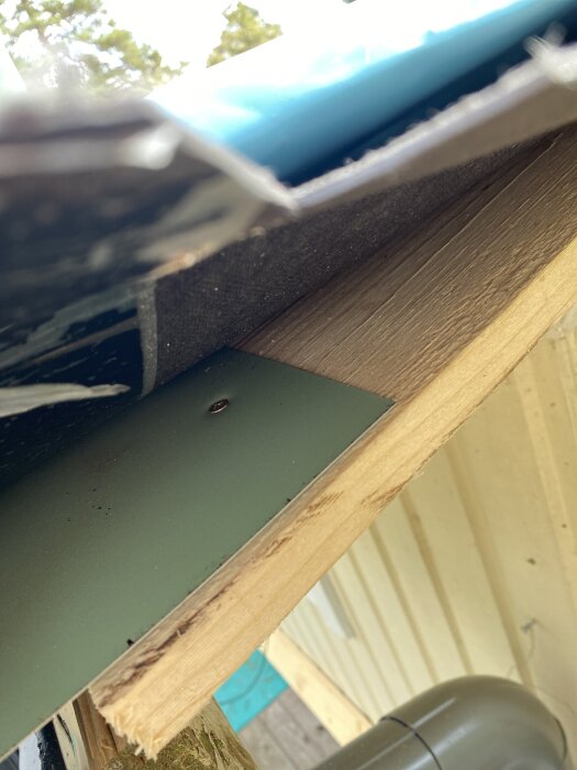 En närbild på takets underkant där vindskivorna är borttagna för målning, med fokus på en träbalk och ett grönt metallstycke.