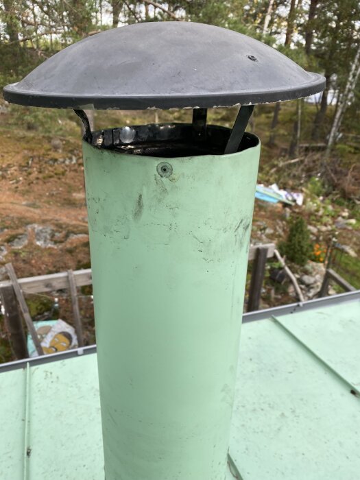Grön skorsten med svart hatt fotograferad utomhus bland träd i bakgrunden.