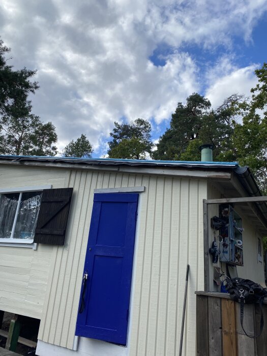 Husvägg med blå dörr och fönster. Taket saknar vindskivor för målning. Skorstenens position har justerats.