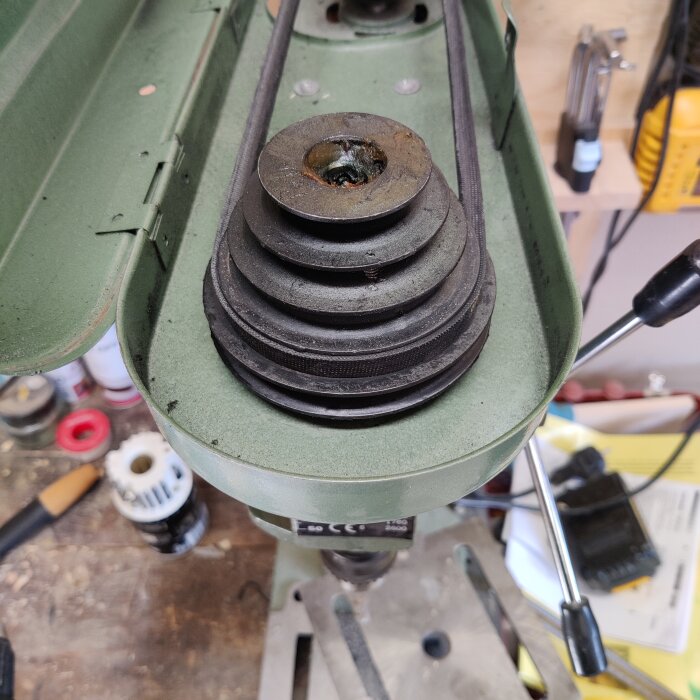 En grön borrmaskin sedd ovanifrån, med en svänghjulsmekanism och remmar synliga. Bakgrunden visar ett verktygsbord med olika verktyg och material.