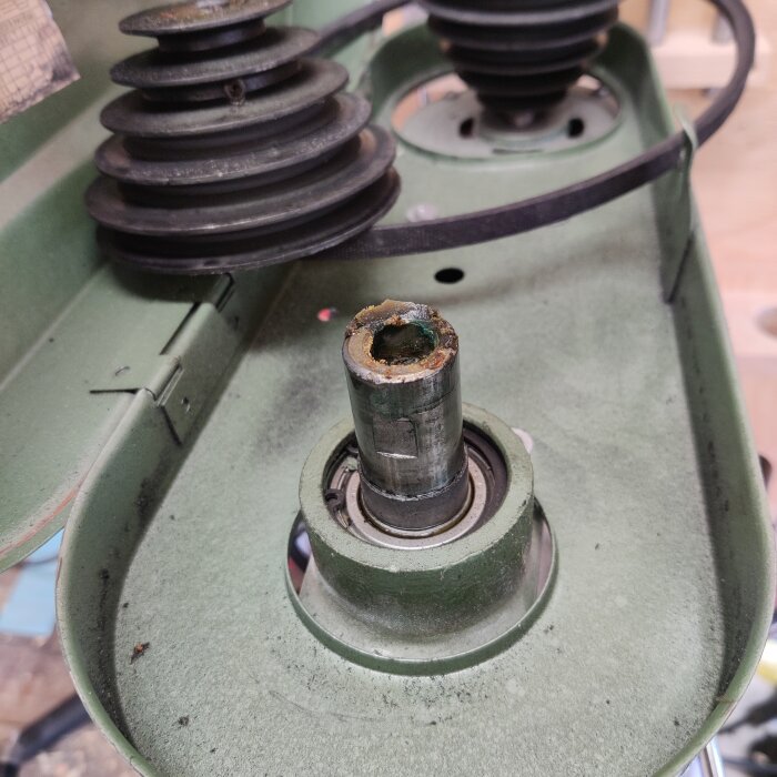 En rostig metallaxel fotograferad ovanifrån, omgiven av en grön metallram med svarta remskivor.