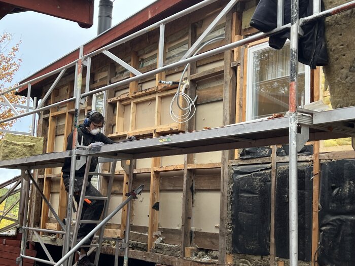 Person arbetar på byggnadsställning med husisolering och fönsterinstallation.