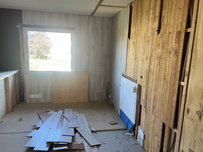 Rum under renovering med staplade träplankor på golvet, exponerade träväggar och ett fönster; förberedelser för att bygga om till barnrum.
