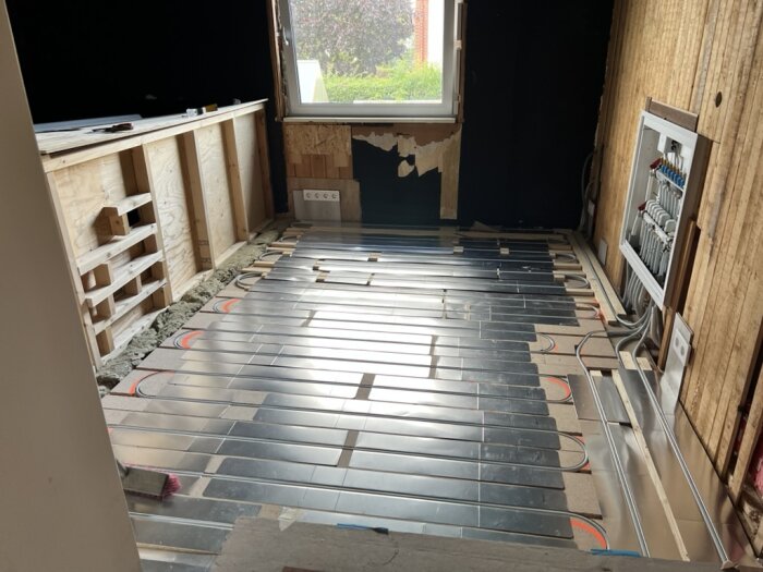 Ett rum under renovering med synliga golvvärmerör och en vägg med exponerad isolering och kablar, samt ett fönster som släpper in ljus.