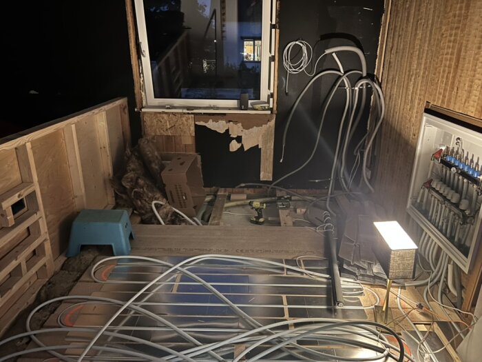 Ett under renovering barnrum med synliga rör och ledningar på golvet, ett öppet fönster och en ljuslykta. Golvvärmesystem ses även på golvet.