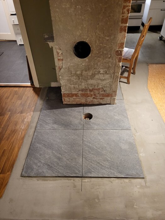 Grått klinkergolv installerat framför putsad murstock med rökkanal och synliga tegelstenar, omgiven av trägolv och grått klinker.