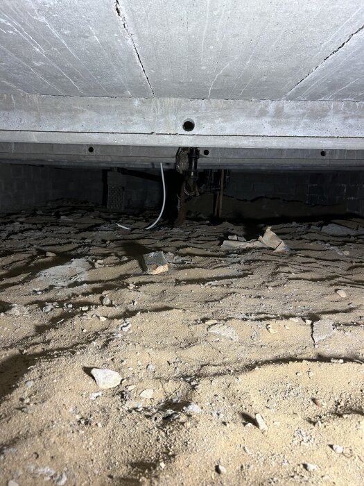 Krypgrund med betongbjälklag och exponerade rör, inklusive vit PEX-rör, över en torr sandig yta.
