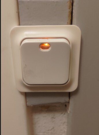 Vit tryckknappsbrytare med en röd glimlampa, monterad på en vägg.