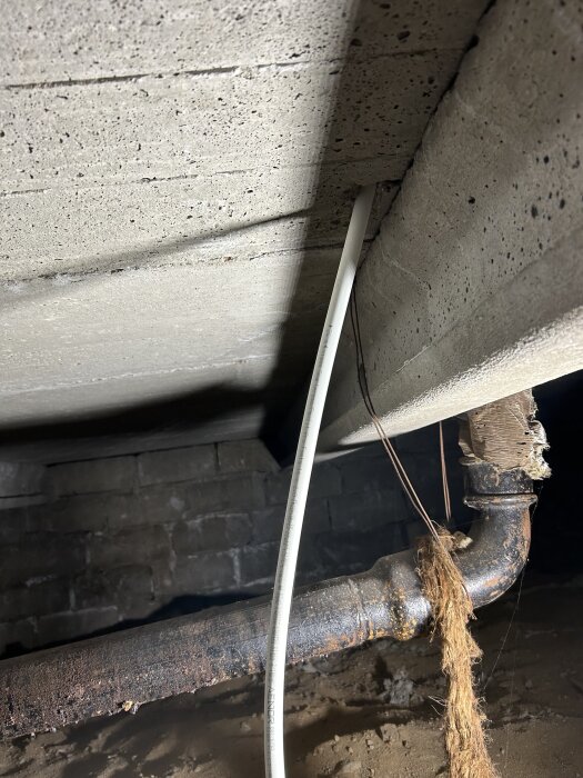 PEX-rör och gammalt avloppsrör i krypgrunden, med betongbjälklag ovanför och murad vägg i bakgrunden.