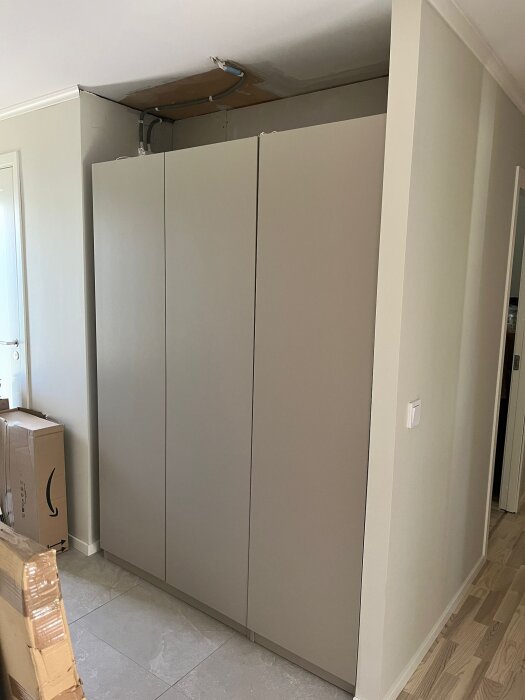 Inbyggd garderob i hall, fast mot väggen. Synlig takpjäs. Kartonger bredvid. Diskussion om byggnation och materialval runt garderoben.