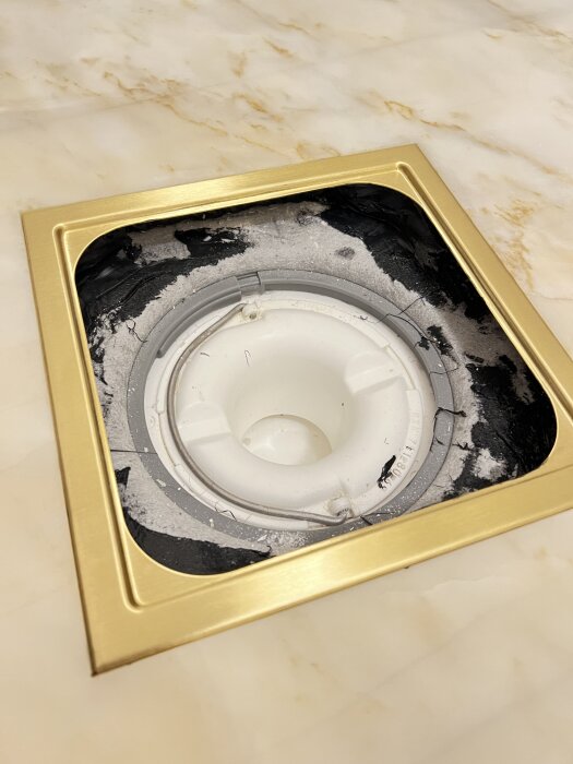 Guldfärgad duschbrunn med osnygg fog på marmorplatta, med svarta partier och ojämnheter runt brunnsöppningen.