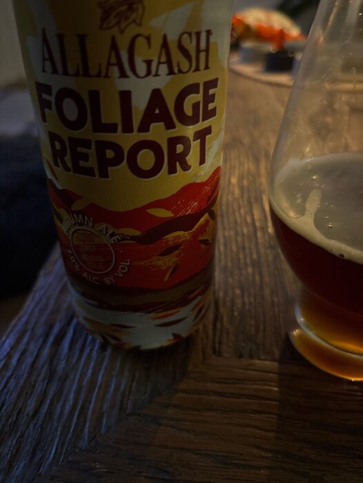 Burk med etiketten "Allagash Foliage Report" och ett glas med mörkt öl på ett träbord.