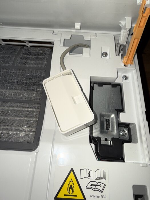 Närbild av insidan av en vit apparat där en panel är borttagen, visar kablage och varningar för R32-kylmedel. Inga synliga knappar.