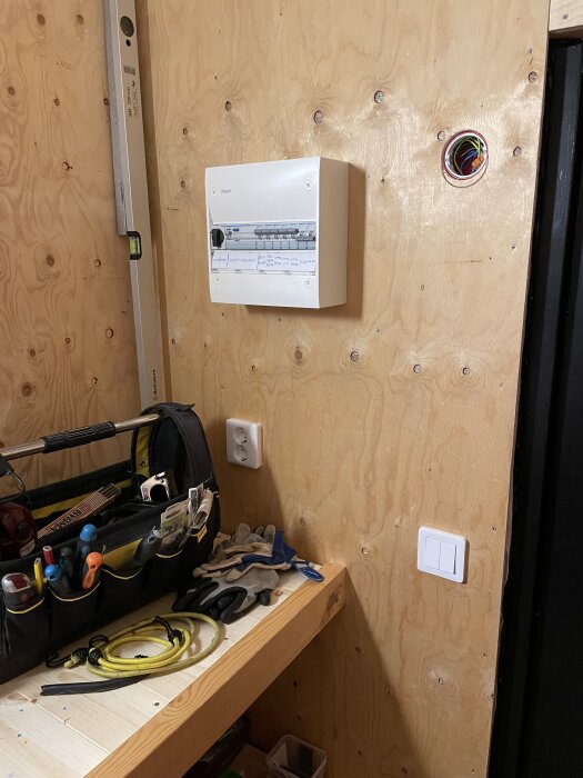 Elektrisk central monterad på plywoodvägg, omgiven av verktygslåda med verktyg och kablar, intill eluttag och strömbrytare.