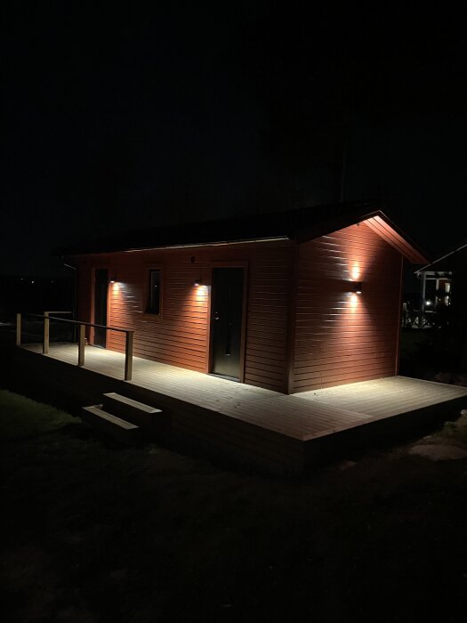 Röd stuga med altan, upplyst i mörkret med utomhusbelysning.